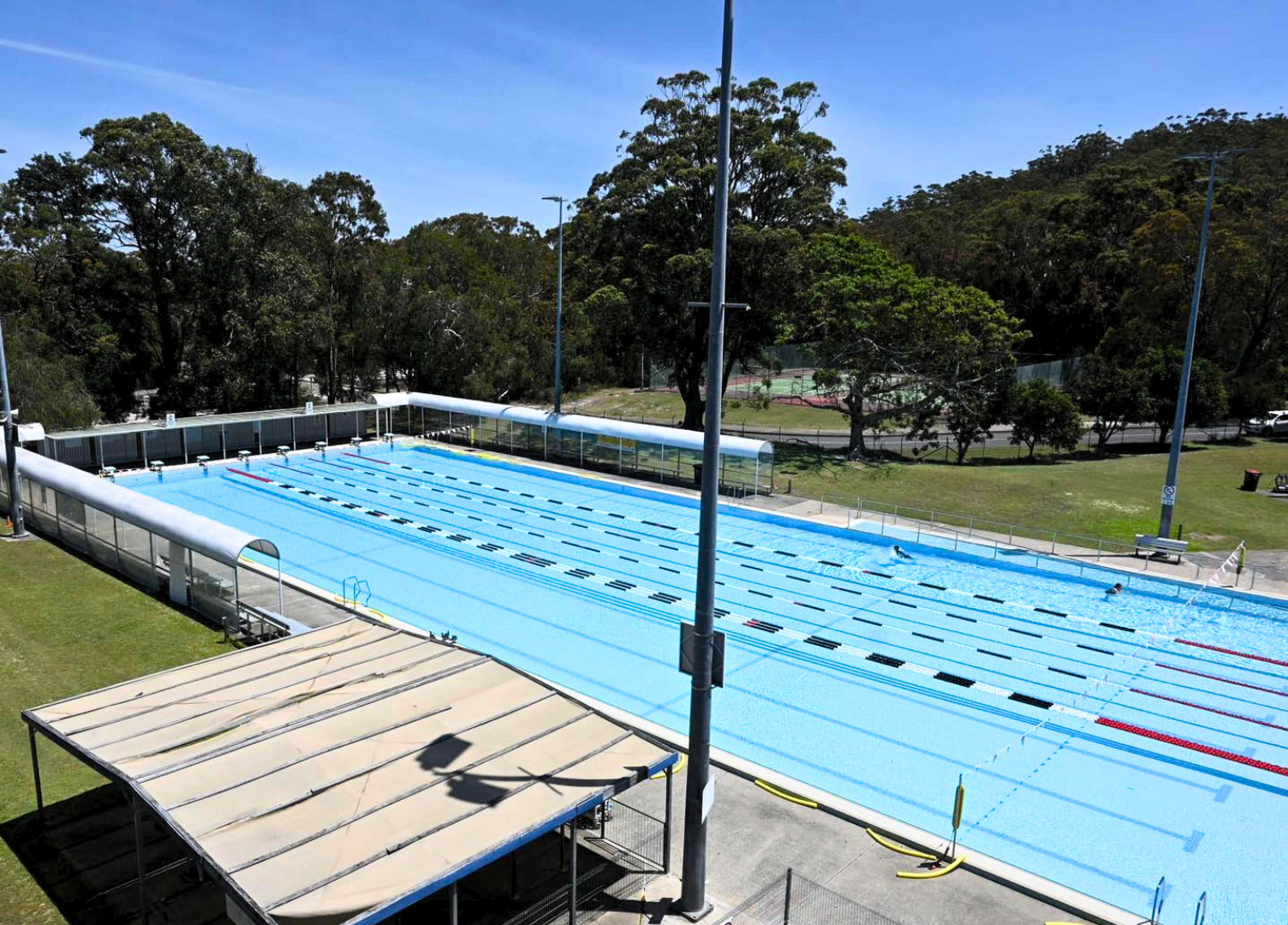 Image shows the 50-metre outdoor heated pool at Tomaree Aquatic Centre. In view is also the roof of the Kiosk and an open lawn area. Clear blue skies.