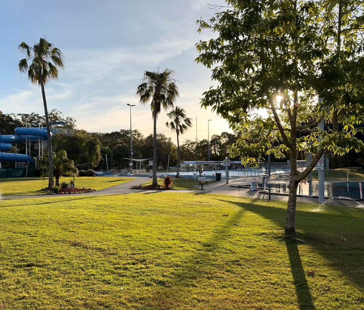 Image shows the grass area at Tomaree Aquatic Centre. The water slide is featured in the background, and the outdoor swimming pool.