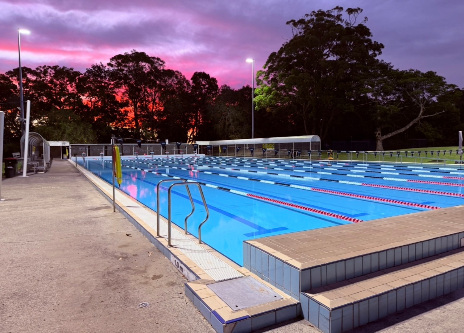 The view of the 50m outdoor heated pool at Tomaree Aquatic Centre in the evening, with purple pink skies. The pool is empty and appears calm.
