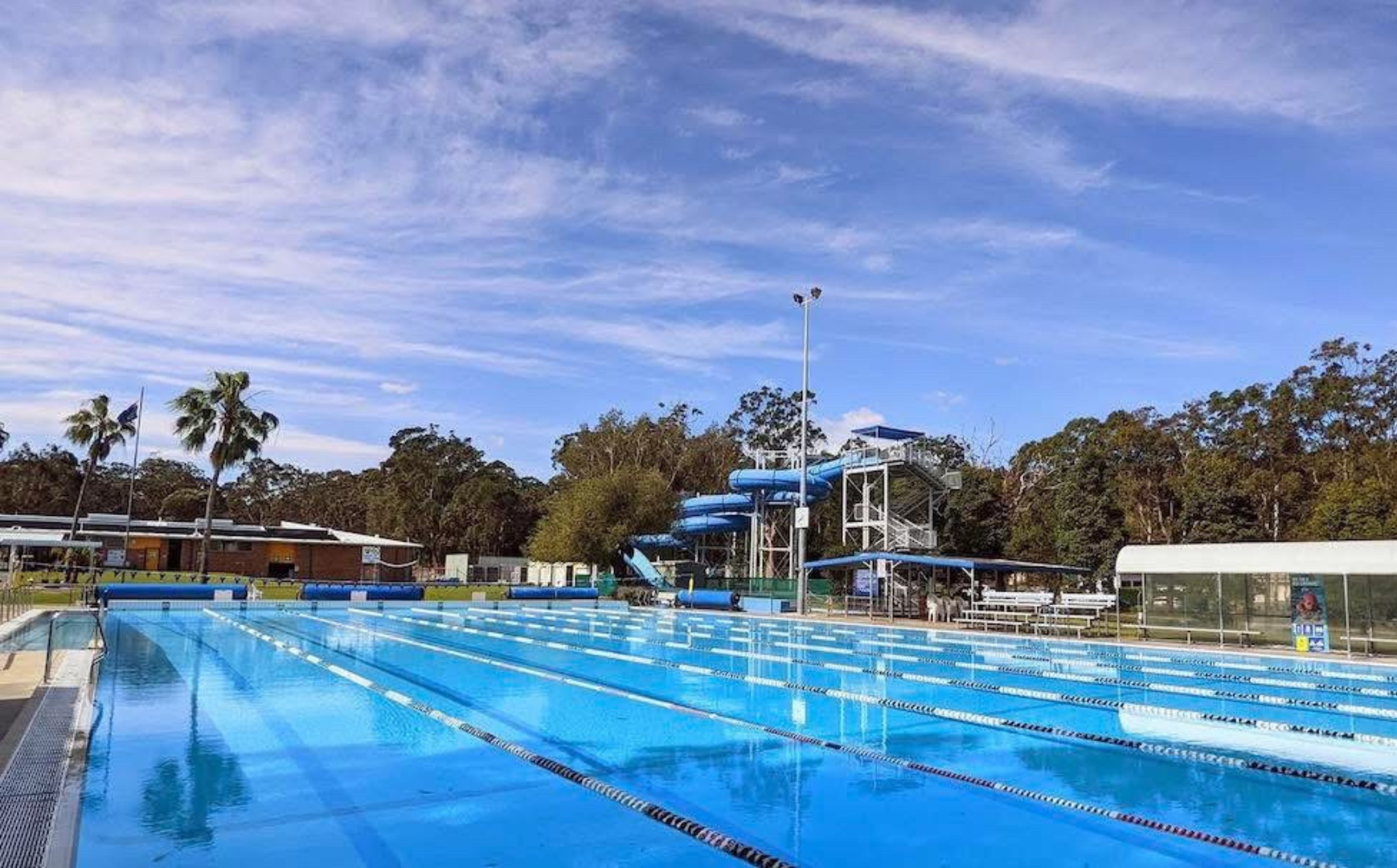 Image shows the grass area at Tomaree Aquatic Centre. The water slide is featured in the background, and the outdoor swimming pool.