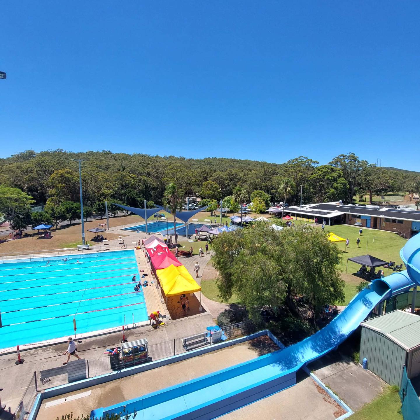 View from above for Tomaree Aquatic Centre - There is the 50m outdoor heated swimming pool and the end of the spiral waterslide. It is a nice sunny day with no clouds and people are enjoying the pool.